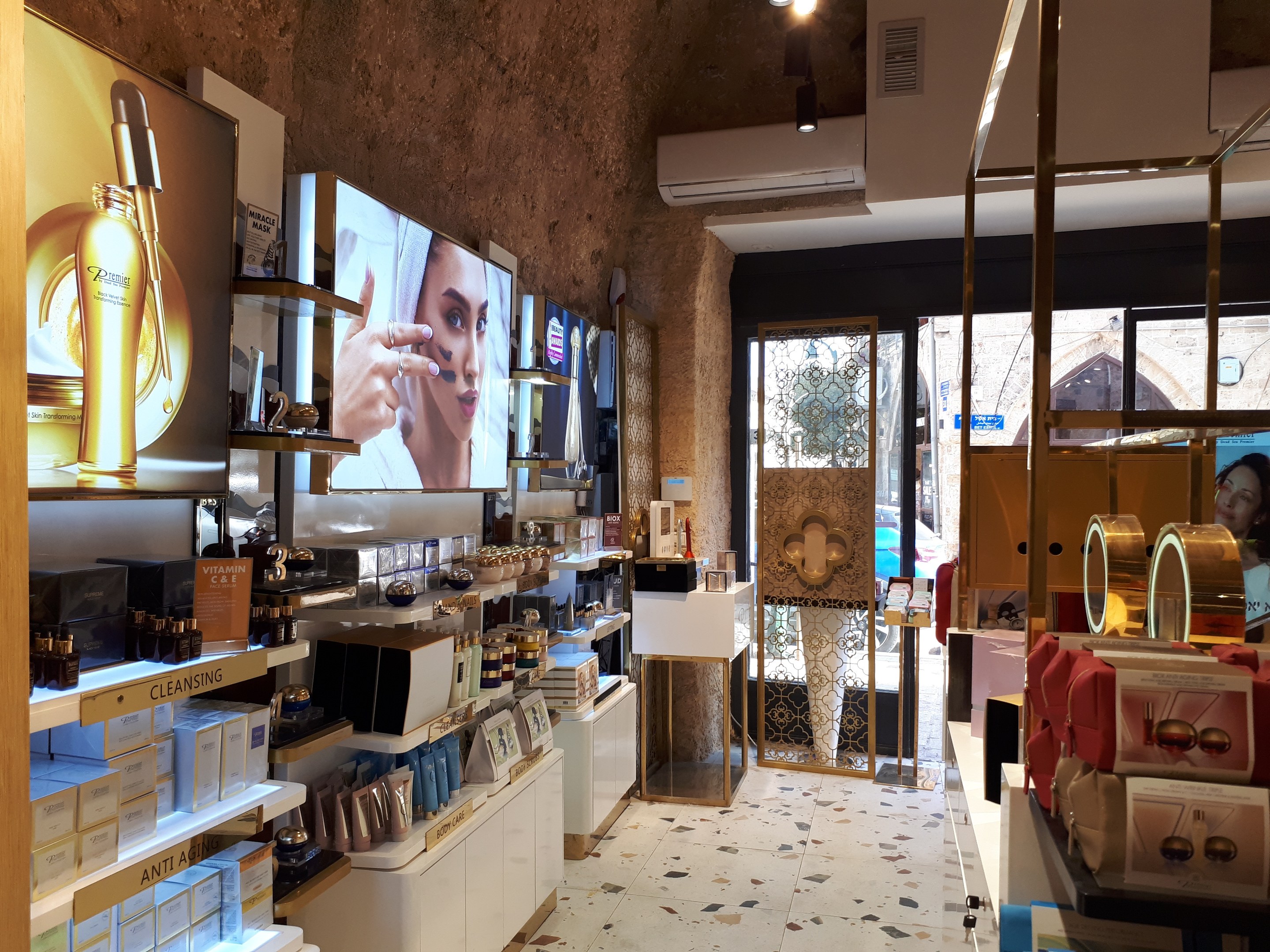 Innenansicht eines Kosmetikgeschäfts mit verschiedenen Produkten auf Gestellen, Wandbildschirmen, Deckenbeleuchtung, einer Klimaanlage und Glastüren, die eine Person, ein Auto und ein Gebäude draußen zeigen.