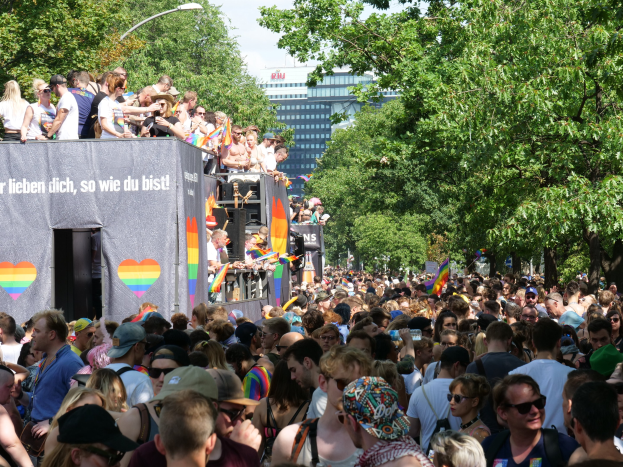 Eine große Menge steht vor einem Lastwagen bei der Christopher Street Day Parade in Berlin, viele tragen Mützen und Schutzbrillen, einige halten Fahnen, mit einem Banner auf dem Lastwagen und Bäumen, Gebäuden und einem Laternenpfahl im Hintergrund bei bewölktem Himmel.