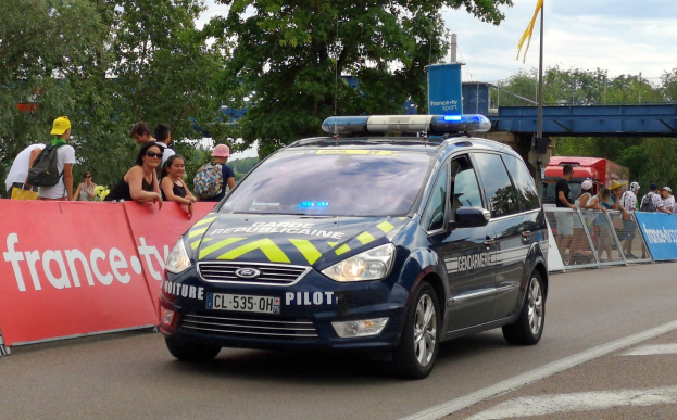 Polizeiauto fährt neben einer Menge mit Schildern, Geländern, Bäumen, einer Brücke, einer Fahne und einem bewölkten Himmel im Hintergrund.