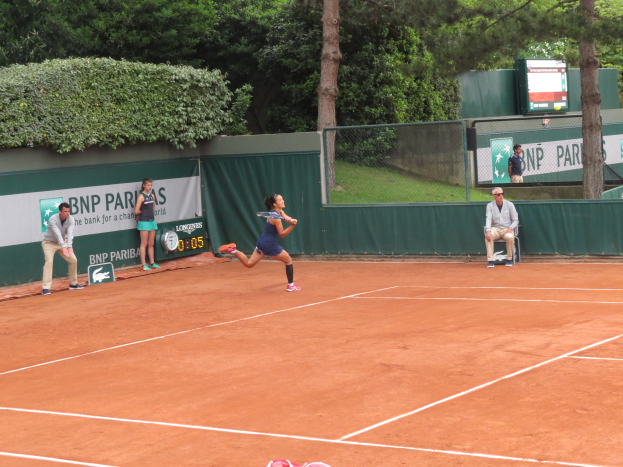 Eine Frau spielt Tennis auf einem Sandplatz, hält einen Schläger und ist von einer Gruppe von Menschen umgeben, darunter eine Person, die rechts von ihr auf einem Stuhl sitzt, sowie ein Zaun, Banner, ein Bildschirm und Bäume im Hintergrund.