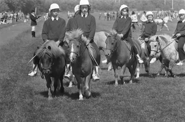 Schwarzes Foto von Kindern mit Helmen auf Pferden, umgeben von einer Menge, Bäumen, Pfählen und einem klaren Himmel.