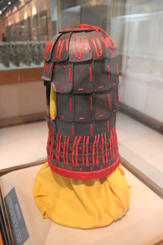 Ein Helm, der in einer Glasvitrine mit einem gelben Tuch darüber ausgestellt ist, begleitet von einer Texttafel auf der linken Seite und Deckenlampen im Hintergrund.