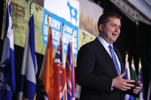 Ein Mann im Anzug spricht an einem Podium mit Mikrofonen, umgeben von Flaggen und einem Bildschirm, wahrscheinlich zu einer Regierungszahlung für die Milchindustrie.