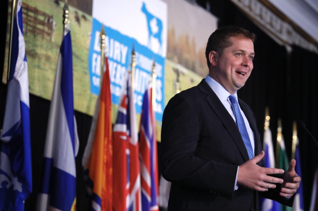 Ein Mann im Anzug spricht an einem Podium mit Mikrofonen, umgeben von Flaggen und einem Bildschirm, wahrscheinlich zu einer Regierungszahlung für die Milchindustrie.