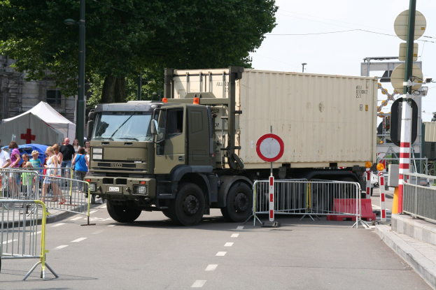 Ein Lkw parkt am Straßenrand, umgeben von Menschen, Absperrungen, Schildern, Straßenlaternen, Strommasten, Stromkabeln, Bäumen, Gebäuden und einem klaren blauen Himmel.