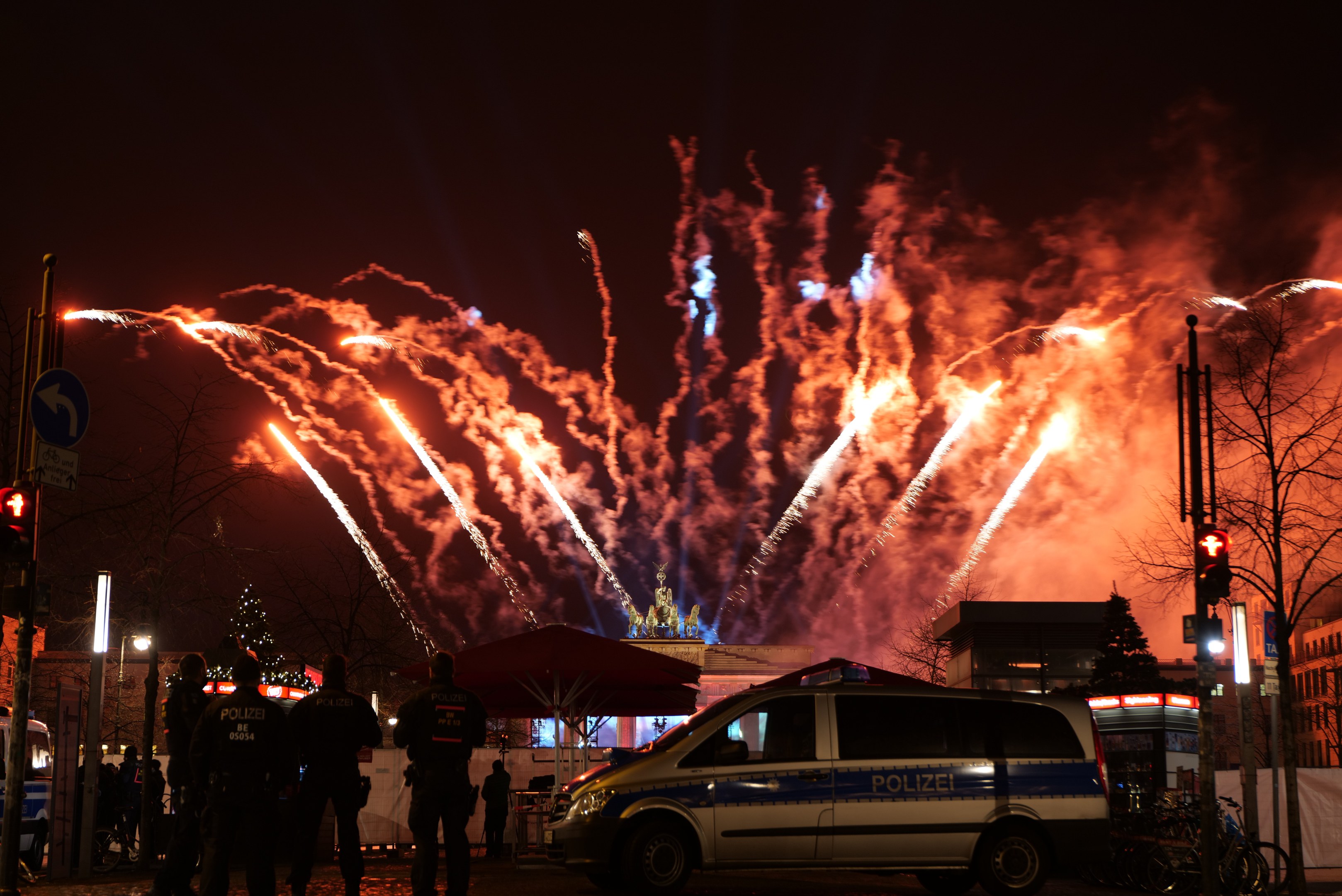 Ein Polizeiauto steht vor einer Menschenmenge, mit beleuchteten Gebäuden und Bäumen im Hintergrund, während farbige Feuerwerke den Himmel erhellen und die Neujahrsfeierlichkeiten in Berlin begleiten.
