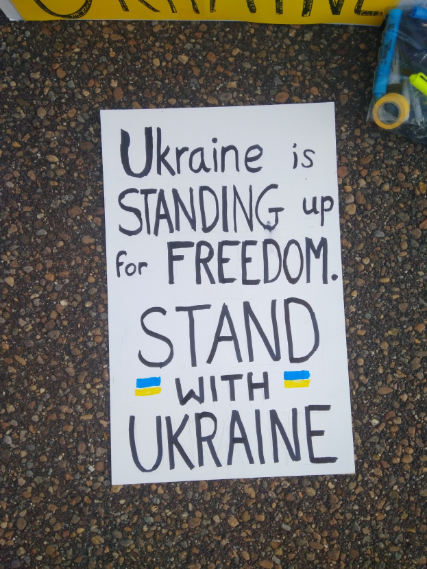 Ein weißer Zettel mit der Aufschrift "Ukraine steht für Freiheit, steht mit Ukraine" liegt vor einer gelben Fahne auf dem Boden. Rechts daneben befindet sich eine Plastikabdeckung mit verschiedenen Gegenständen.