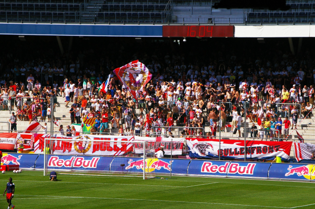 Ein Fußballspiel in einem Stadion mit Spielern auf dem Feld, einem Torpfosten mit Netz, Bannern, einem Metallzaun, einer Anzeigetafel, einem Displaybildschirm und einem Dach mit Deckenbeleuchtung.