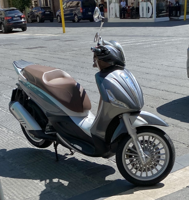 Ein Roller mit einem Helm darauf auf einer Straße geparkt, mit Gebäuden, Bäumen, Polen, Menschen, Stühlen, Tischen und anderen Objekten im Hintergrund.