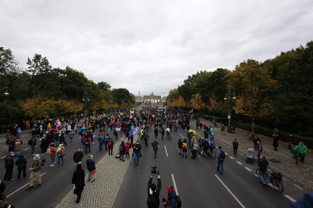 Eine große Gruppe von Menschen marschiert eine von Bäumen gesäumte Straße in Berlin entlang, mit Kameras bewaffnet, mit einem Gebäude und einem klaren Himmel im Hintergrund.