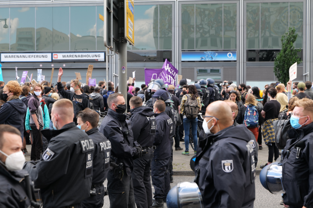 Eine große Gruppe von Menschen steht vor einem Gebäude, einige halten Schilder und tragen Helme, mit einem Schildständer im Vordergrund und einem Baum im Hintergrund, was auf eine Protestaktion hindeutet.
