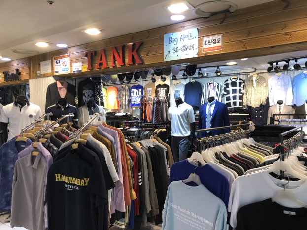 Ein modernes Bekleidungsgeschäft mit zahlreichen Shirts an den Wänden, Schaufensterpuppen, die aktuelle Outfits präsentieren, beleuchteter Decke und Informationsschilder an den Wänden.