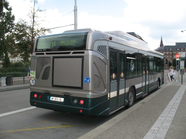 Eine Stadtbus fährt auf einer von hohen Gebäuden gesäumten Straße, mit Fußgängern auf einem Gehweg auf der rechten Seite und Schildern auf der rechten Seite, und Bäumen, Gebäuden und Himmel im Hintergrund.