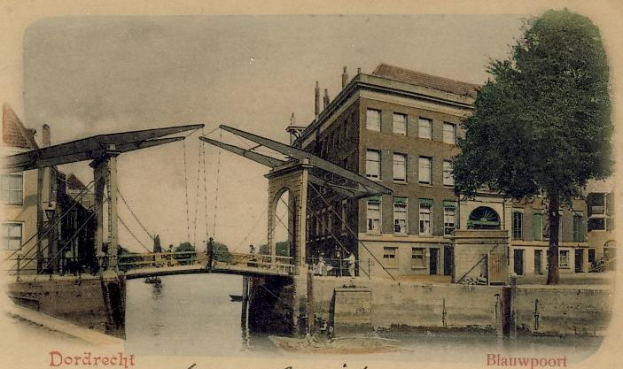 Altes Schwarz-Weiß-Foto einer Brücke unter Bau, die einen Fluss überspannt, mit Gebäuden, Bäumen und einem klaren Himmel im Hintergrund; Werkzeuge und Materialien sind auf der Brückenfläche sichtbar, und Text erscheint unten.