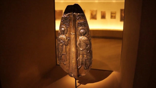 Ein byzantinisches Metallobjekt in einer Glasvitrine, beleuchtet und von gerahmten Fotos umgeben in einer Museumsausstellung.
