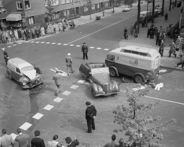 Schwarze und Weiße Szenerie eines Autounfalls an einer Straßenecke mit mehreren Fahrzeugen, darunter ein Lieferwagen, und einer Gruppe von Menschen, mit Gebäuden, Bäumen, Masten und Texttafeln im Hintergrund.