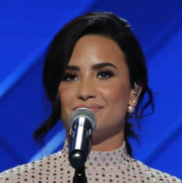 Demi Lovato steht selbstbewusst vor einem Mikrofon bei den Grammys, trägt ein schwarzes Kleid und eine glatte Bob-Frisur, vor einem blauen Hintergrund.