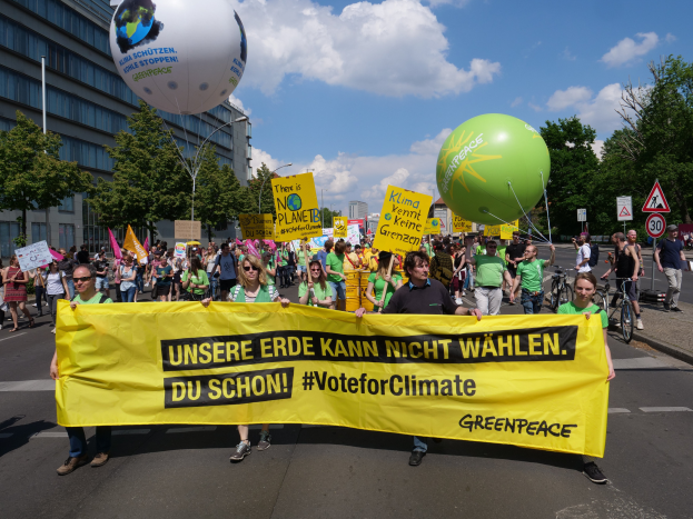 Eine Gruppe von Menschen marschiert mit einem gelben "Wähle für das Klima"-Schild die Straße entlang, mit Ballons, Schildern, Fahrrädern, Bäumen, Laternenpfählen und Gebäuden im Hintergrund unter einem klaren blauen Himmel.