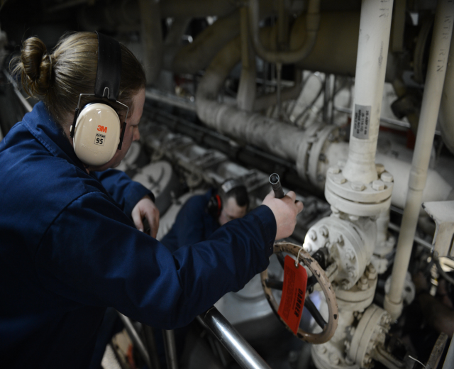Zwei Frauen in blauen Uniformen arbeiten an einer Maschine in einer Öl- und Gasindustrieumgebung, wobei eine ein Objekt hält und Kopfhörer trägt und Rohre im Hintergrund sichtbar sind.