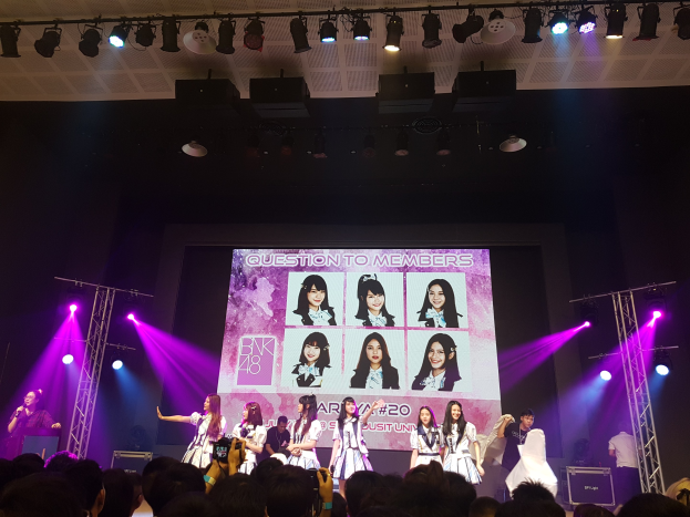 Eine Gruppe von Menschen auf einer hell erleuchteten Bühne, wobei einer ein Mikrofon hält und ein anderer ein Instrument spielt, die vor einem Publikum auftreten, während im Hintergrund ein großes Display Bilder der K-Pop-Gruppe BK48 zeigt.