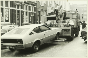 Ein Schwarz-Weiß-Foto eines Autos, das von einem Abschleppwagen auf einer Straße mit anderen Fahrzeugen, einer Person in der Nähe und Gebäuden mit Fenstern im Hintergrund geschleppt wird.