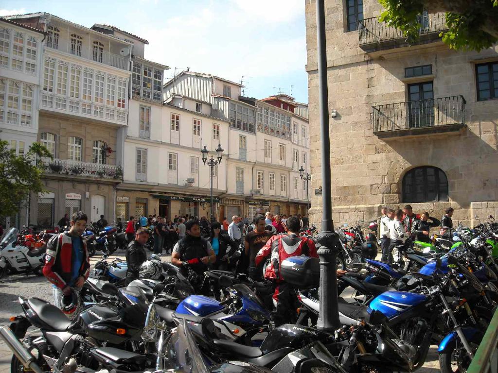 Ein cremefarbenes Gebäude mit zahlreichen Fenstern, neben einem Backsteingebäude mit einer Metallbalustrade, mit mehreren geparkten Fahrrädern und Menschen, die vor dem Gebäude sprechen.