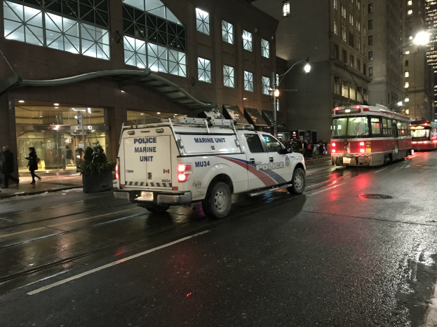 Ein Polizei-Lkw parkt nachts am Straßenrand, mit Fahrzeugen auf der Straße, Fußgängern auf dem Gehweg, Topfpflanzen, Straßenlaternen, Gebäuden mit Fenstern und einer bewölkten Nacht.