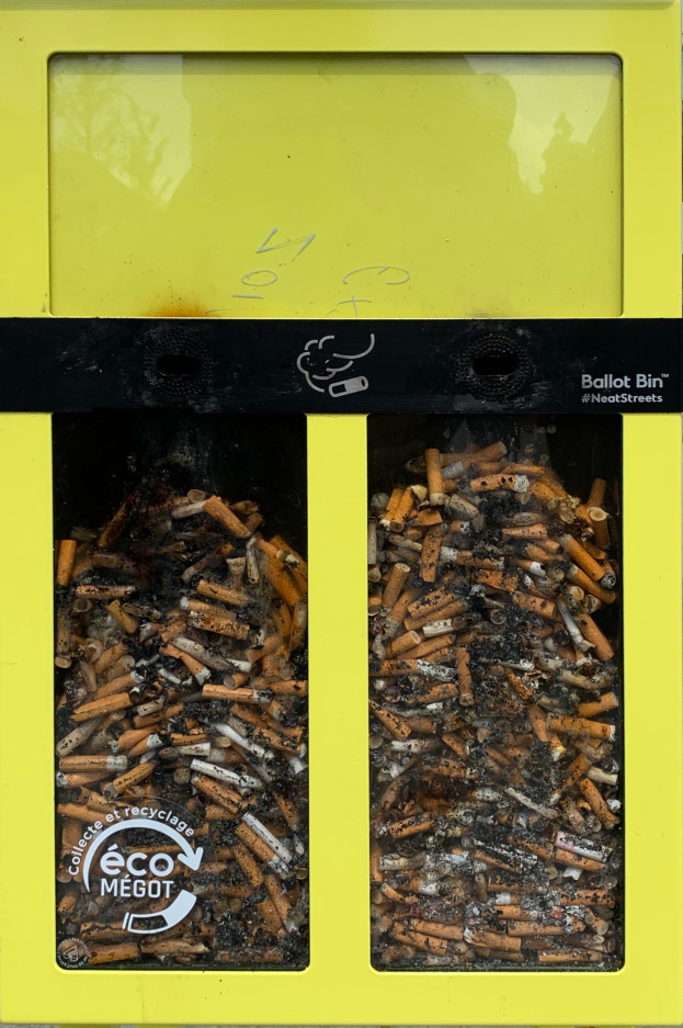 Gelber Recycling-Eimer voller Zigarettenstöpsel auf einem Gehweg, mit Text auf dem Eimer.