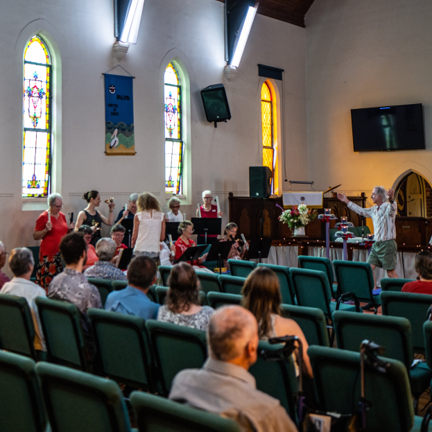 Eine Gruppe von Menschen sitzt auf Stühlen in einer Kirche, ein Mann steht vorne mit einem Mikrofon in der Hand, umgeben von Musikinstrumenten, einem Tisch mit einer Blumenvase, einem Lautsprecher, einer Wandbanner mit Text, Fenstern und Deckenleuchten.