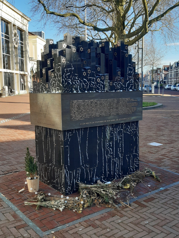 Denkmal für Holocaust-Opfer in einem Stadtplatz, mit einem Baum davor, einem Topfgewächs, verstreuten trockenen Blättern und Gebäuden im Hintergrund.