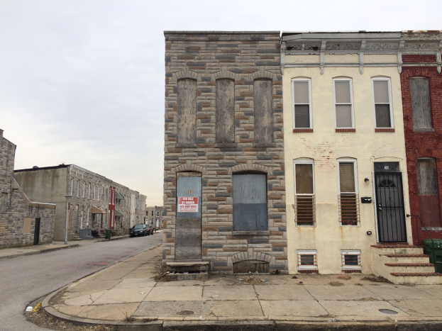 Ein altes, baufälliges Gebäude an einer Straßenecke mit zerbrochenen Fenstern, bröckelnden Wänden, vorbeifahrenden Fahrzeugen, Mülltonnen entlang des Bürgersteigs und einem klaren blauen Himmel im Hintergrund.