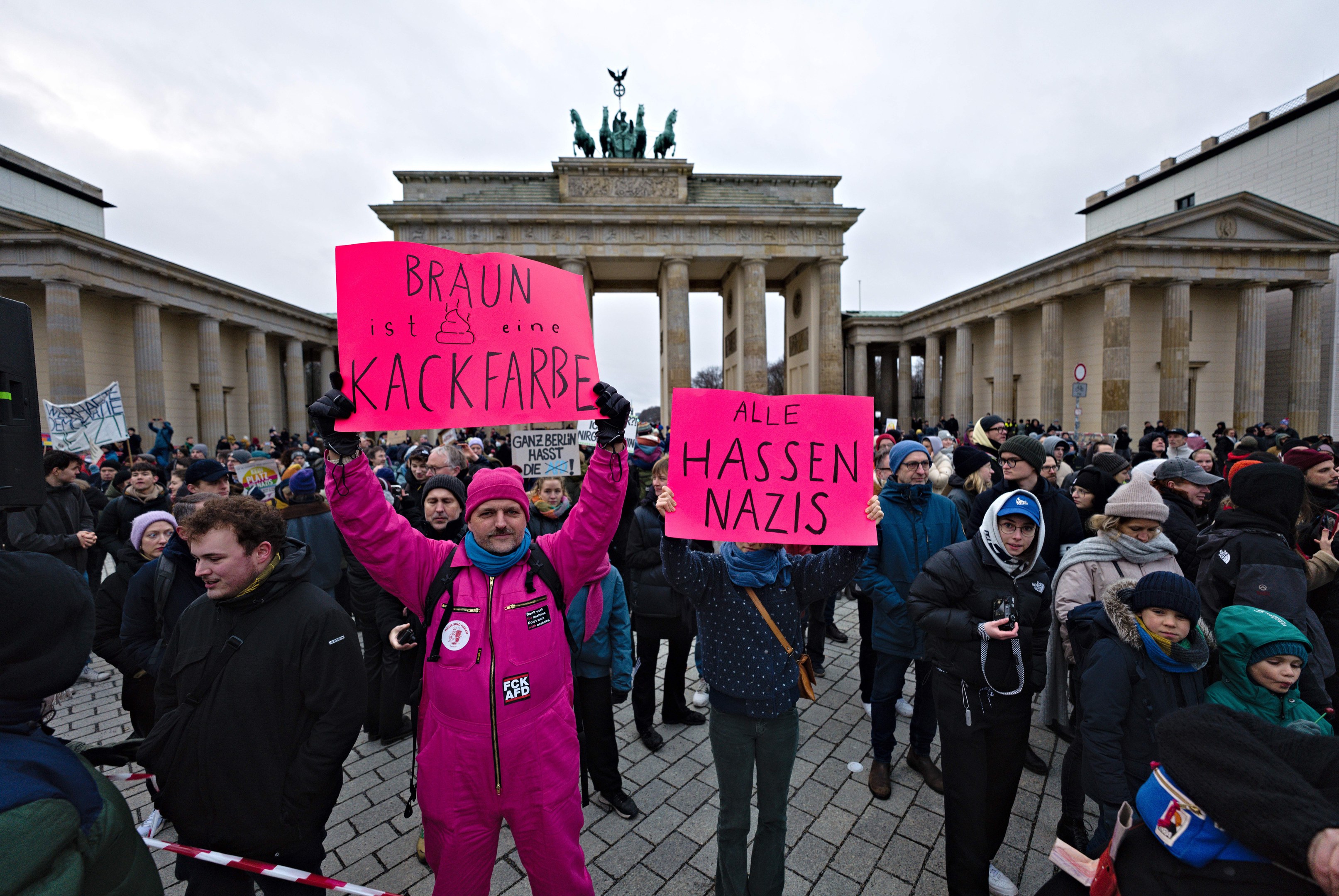 Gruppe von Menschen protestiert vor dem Brandenburger Tor, hält Schilder gegen die Anti-Nazis-Bewegung der deutschen Regierung, mit Gebäuden und Statuen im Hintergrund unter einem klaren blauen Himmel.