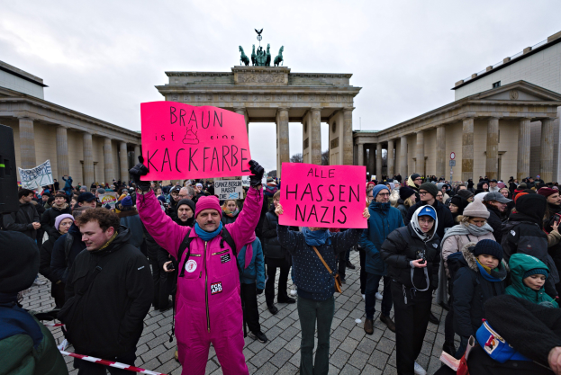 Gruppe von Menschen protestiert vor dem Brandenburger Tor, hält Schilder gegen die Anti-Nazis-Bewegung der deutschen Regierung, mit Gebäuden und Statuen im Hintergrund unter einem klaren blauen Himmel.