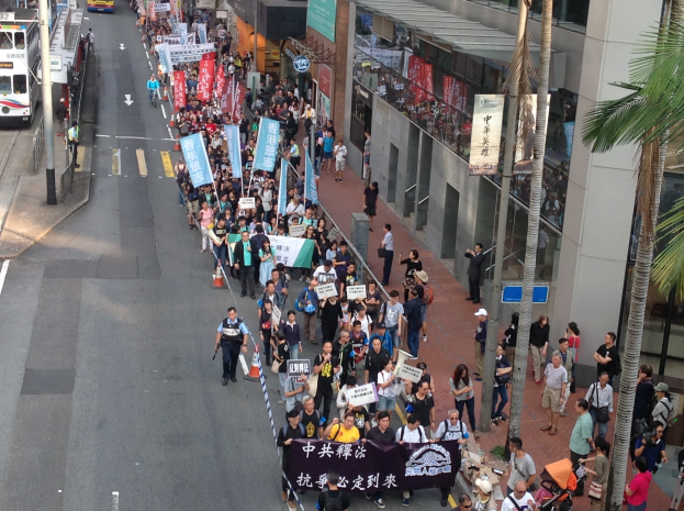 Eine große Gruppe von Menschen marschiert auf einer Stadtstraße und hält Schilder und Banner hoch, mit Bäumen, Gebäuden und Fahrzeugen im Hintergrund.