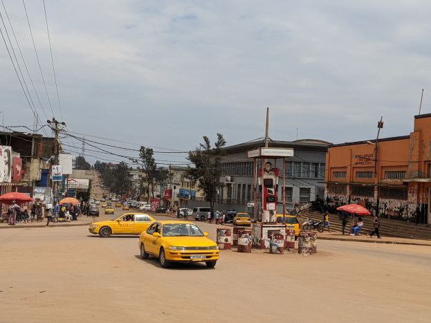 Ein gelbes Taxi fährt eine belebte Stadtstraße mit Gebäuden an den Seiten hinunter, während Fußgänger mit Schirmen auf dem Gehweg darunter unter bewölktem Himmel gehen.