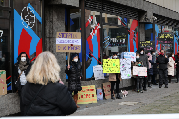 Eine Gruppe maskierter Menschen protestiert vor einem Glasgebäude, hält Schilder und Plakate hoch, mit Kameras und einem Hund dabei.