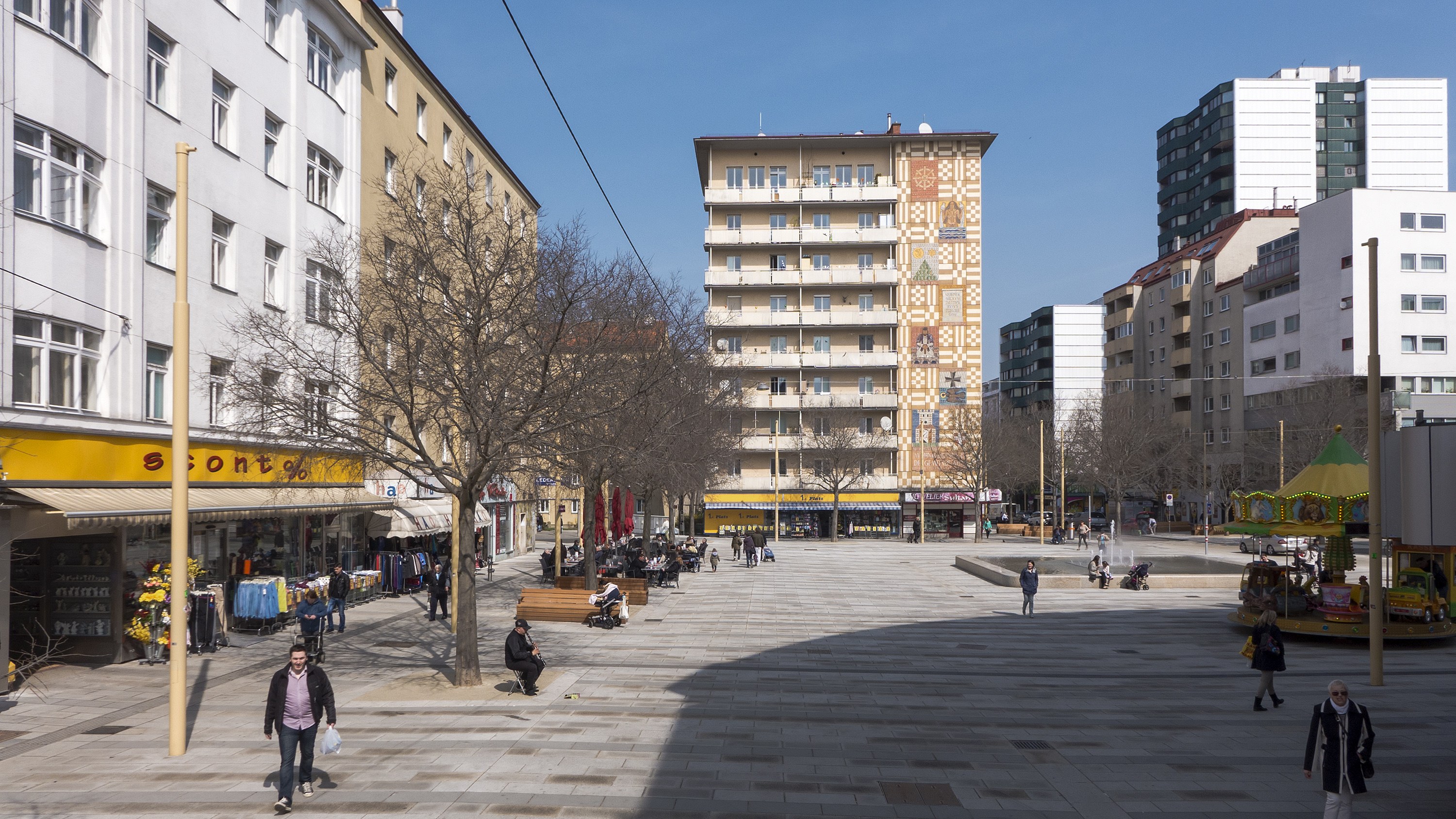 Ein Stadtplatz mit Fußgängern, Bänken, Bäumen, Gebäuden, einer Karussell und Stromleitungen gegen einen sichtbaren Himmel.