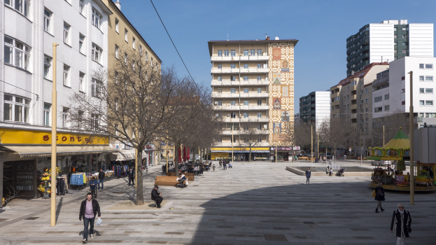 Ein Stadtplatz mit Fußgängern, Bänken, Bäumen, Gebäuden, einer Karussell und Stromleitungen gegen einen sichtbaren Himmel.