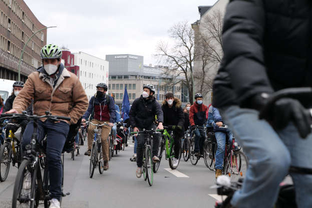 Eine Gruppe von Menschen in Helmen und Handschuhen, die Fahrräder auf einer von Bäumen gesäumten Straße in Berlin fahren, mit Gebäuden und einem geparkten Fahrzeug im Hintergrund.