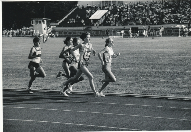 Eine Gruppe von Frauen, die auf einer Laufbahn in einem Stadion laufen, mit einer Zuschauermenge im Hintergrund, in Schwarz-Weiß.