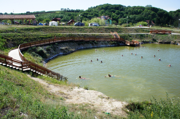 Gruppe von Menschen, die in einem Gewässer schwimmen, mit einer Brücke, Treppen, üppiger Vegetation, Hütten, Fahrzeugen, Pfählen und einem klaren blauen Himmel im Hintergrund.