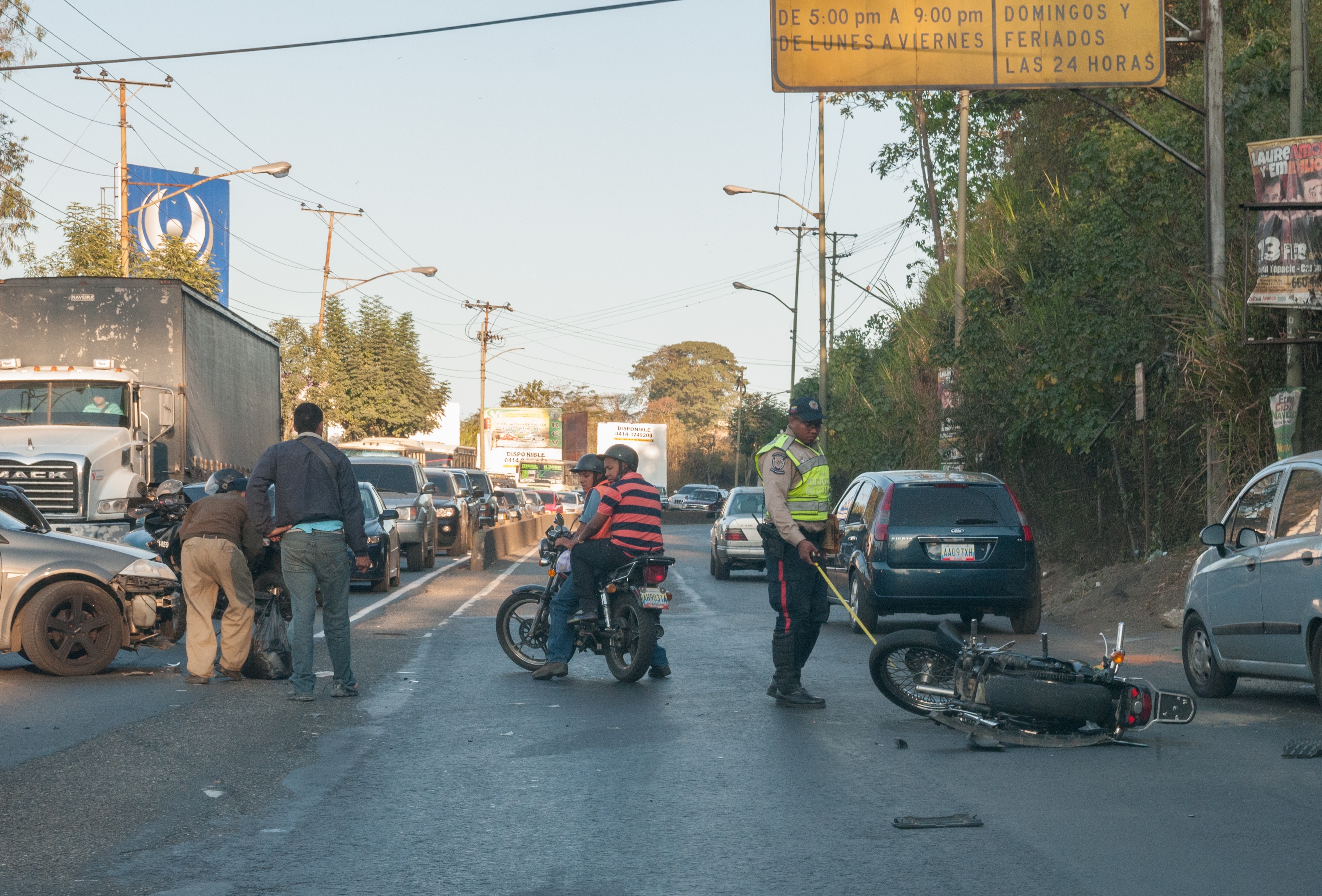 Eine Gruppe von Menschen steht um ein verunglücktes Motorrad auf der Straße herum, umgeben von mehreren Fahrzeugen, einschließlich eines Lastwagens, und einer Hintergrundlandschaft aus Bäumen, Pfählen, Lampen, Schildern und Himmel.