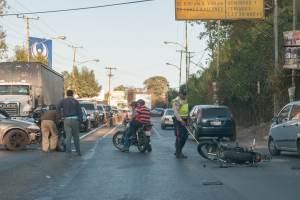Eine Gruppe von Menschen steht um ein verunglücktes Motorrad auf der Straße herum, umgeben von mehreren Fahrzeugen, einschließlich eines Lastwagens, und einer Hintergrundlandschaft aus Bäumen, Pfählen, Lampen, Schildern und Himmel.