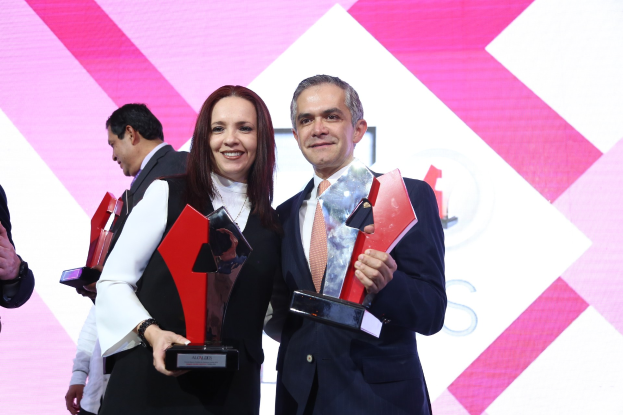 Ein Mann und eine Frau stehen nebeneinander auf einer B├╝hne, jeder h├Ąlt eine Troph├Ąe und l├Ąchelt, mit einem Bildschirm im Hintergrund.