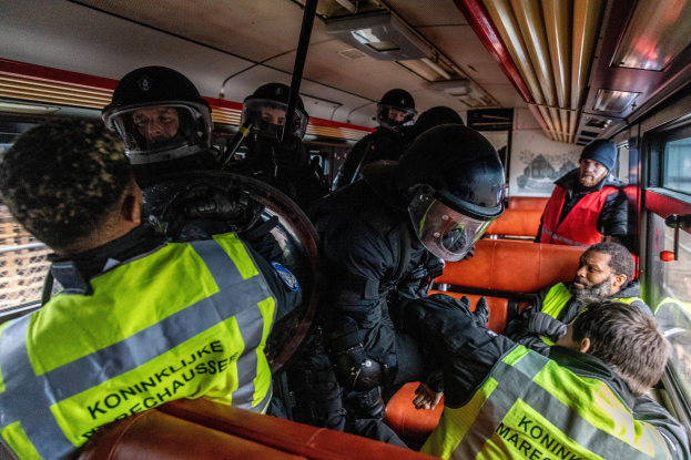 Eine Gruppe von Polizisten in Einsatzausrüstung, einige mit Helmen und Jacken, befindet sich auf einem Bus mit sichtbaren Fenstern und umgibt eine Person, die in der Mitte sitzt, mit einem Plakat im Hintergrund.