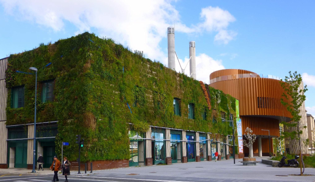 Ein großes Gebäude mit einer grünen Wand an seiner Seite, umgeben von Straßeninfrastruktur, Fahrzeugen, Fußgängern, Bäumen und einem bewölkten Himmel, als Teil des Green Building Projects.