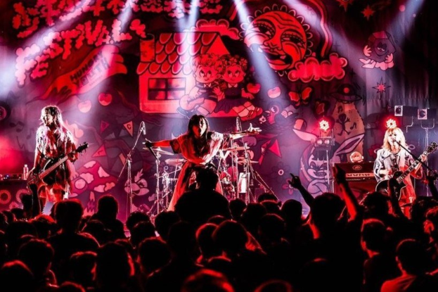 Eine Live-Aufführung der japanischen Rockband Korn auf der Bühne, mit Bandmitgliedern, die Gitarren spielen und in Mikrofone singen, begleitet von Scheinwerfern und einem Gemälde im Hintergrund, während das Publikum von unten zusieht.