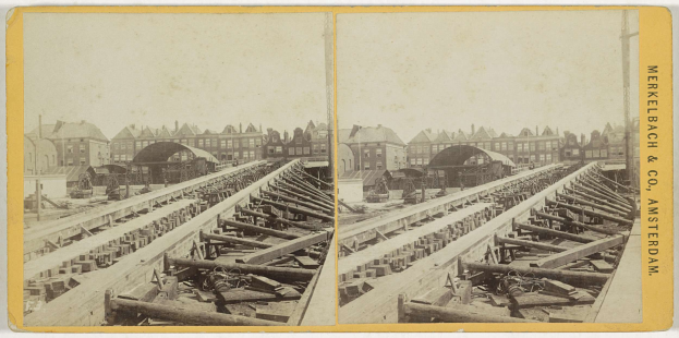 Schwarz-weiß-Foto einer Bahnschiene mit Gebäuden im Hintergrund, Text 'Merkelbach & Co, Co. Amsterdam' rechts und verstreuten Holzgegenständen im Vordergrund.