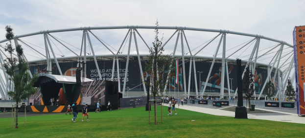 Olympiastadion in London mit Menschen, einem Baum im Vordergrund, Bannern am Gebäude und bewölktem Himmel.