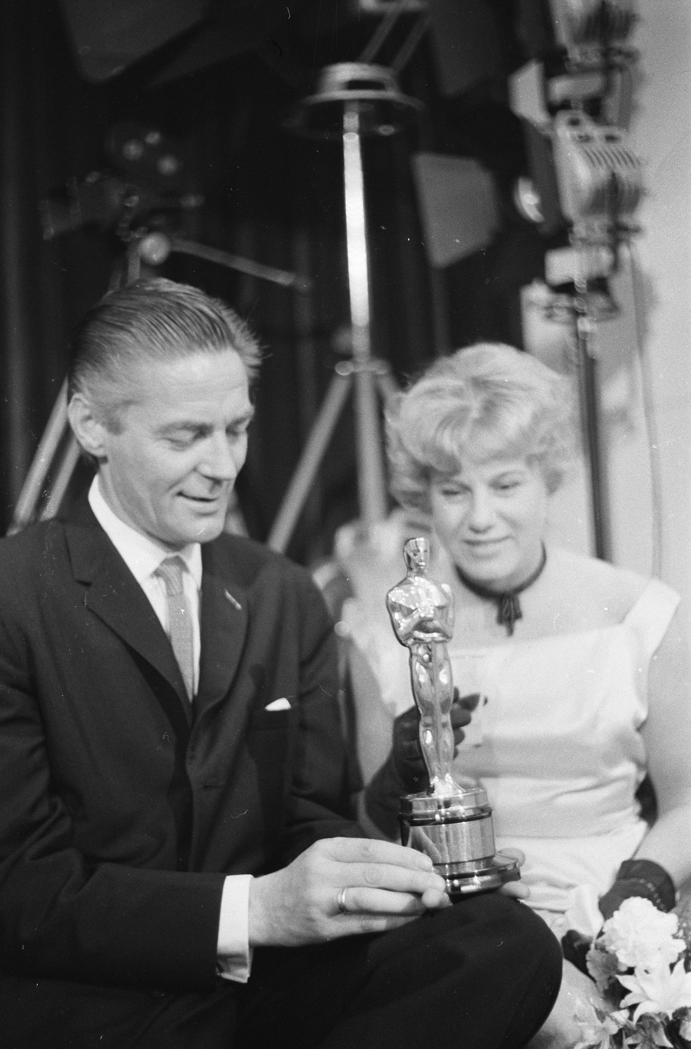 Ein Mann in einem schwarzen Blazer, weißem Hemd und Krawatte hält eine Oscar-Statue in der Hand und sitzt neben einer Frau, die einen Blumenstrauß hält, mit Lichtern im Hintergrund, alles in Schwarz-Weiß.